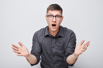 Fototapeta premium Surprised young man with glasses hands raised and mouth open in shock against a white background.