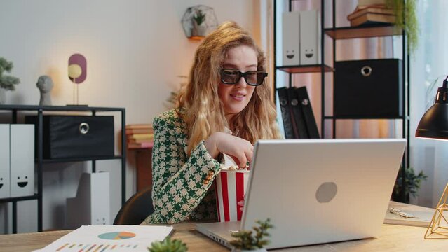 Caucasian businesswoman freelancer taking break from work wearing 3D glasses eating popcorn and watching movie on laptop. Smiling blond girl sitting at home office desk having snacks during break