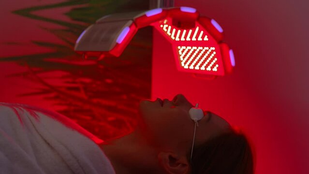 Tracking shot of female patient undergoes red LED light or RLT therapy for skin rejuvenation at modern luxury aesthetic clinic. Woman client having non-invasive type of phototherapy, slow motion.