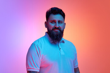 Calm bearded man looking at camera in neon light against vibrant gradient studio background. Male...