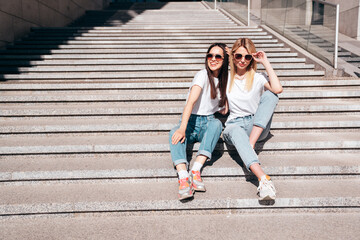 Two young beautiful smiling hipster female in trendy summer white t-shirt and jeans clothes. Carefree women posing in street. Positive models having fun. Cheerful and happy. In sunglasses