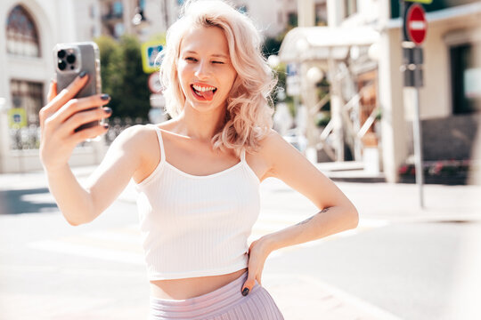 Young beautiful smiling blond hipster woman in trendy summer clothes. Carefree female posing in the street at sunny day. Positive model outdoors at sunset. Cheerful and happy. Takes selfie photos