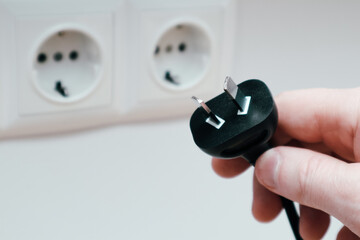 A man's hand holds an electrical plug next to the sockets on the white wall. Different standards of...