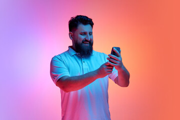 Portrait of young man holding smartphone and smiling lookin to screen in neon light against vibrant...