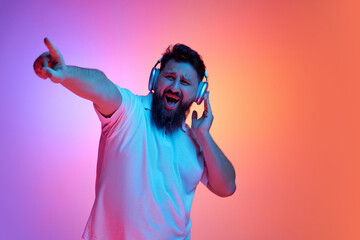 Joyful young man listening music in headphones and dancing in neon light against vibrant gradient studio background. Concept of human emotions, self-expression, beauty and fashion, fun and joy. Ad