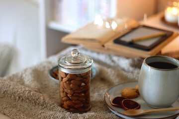 Obraz na płótnie Canvas Cup of tea or coffee, cookies, books, glasses, e-reader, almonds, ball of yarn, knitting needles and lit candles on the table. Hygge at home. Selective focus.