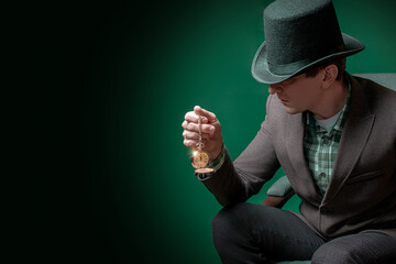 Stylish gentleman in a bowler hat and a cane sits in a chair and looks at an antique watch