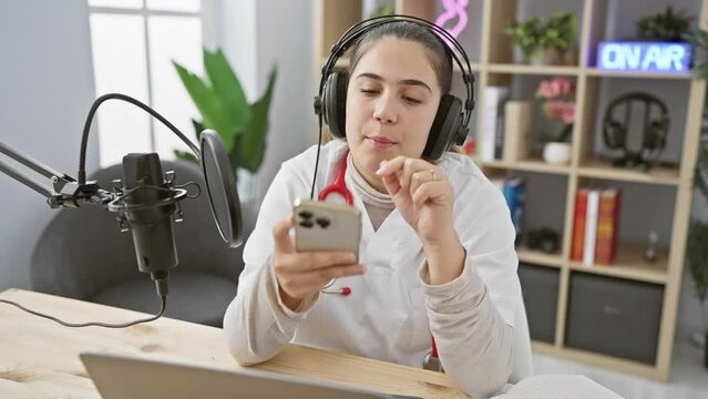 A hispanic woman with headphones in a studio gestures while holding a smartphone, microphone, and 'on air' sign visible.