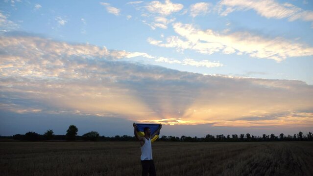Young man stands on barley meadow with raised over head flag of Ukraine at sunset. Ukrainian sportsman looking into camera with lifted national banner at sunrise. Victory against russian aggression
