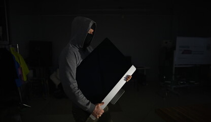 Portrait of man wearing grey hoodie and black mask. Male thief standing in dark room and holding...