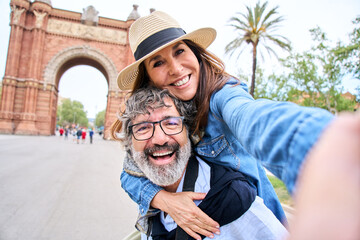 A senior gray hair man wearing a sun hat is giving a piggyback ride to a mature woman in a happy...