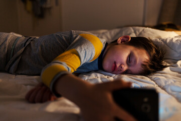 A carefree boy is lying in bed at home, holding a smartphone in his hand while sleeping.