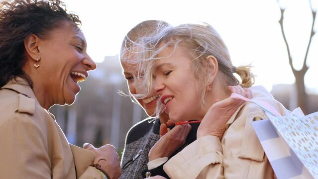 Group gathering of middle aged excited women together chatting and laughing standing on sunny day. Three multiracial friends showing off their outdoor shopping bags. Cheerful female sales and bargains