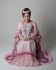 Fun length portrait of female model wearing an opulent pink gown, costume of a historical French baroque nobility, style of Marie Antoinette. Sitting pose on throne. Studio background.