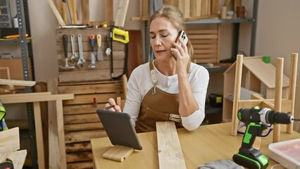 A mature woman carpenter in a workshop multitasks, using a tablet and phone while surrounded by wooden furniture and tools. - Powered by Adobe
