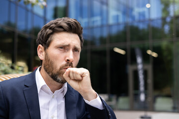 Close-up photo of a young sick man in a business suit who is standing outside an office building and coughing, covering his mouth with his hand