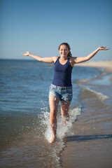 A young barefoot girl is happily running along the ocean shore. She is smiling broadly, her hair is pulled back in a ponytail, she is wearing denim shorts and a blue T-shirt