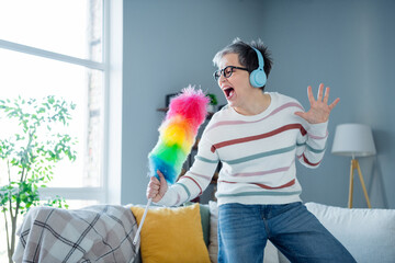 Photo of carefree positive aged person enjoy cleaning listen music headphones sing duster bright...