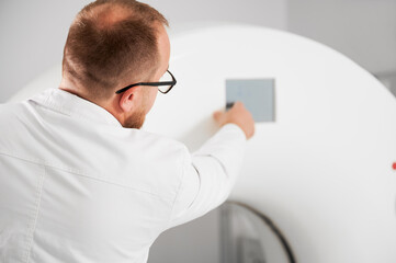 Medical computed tomography or MRI scanner. Back view of male radiologist presses MRI button to...