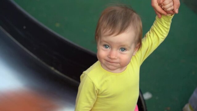 Mother helps little baby girl slide down slide on playground in summer outdoors. Childrens leisure and active games