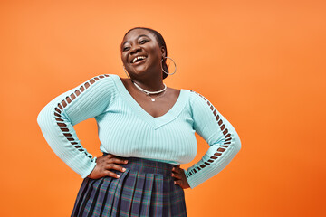 cheerful plus size african american woman in blue long sleeve posing with hands on hips on orange