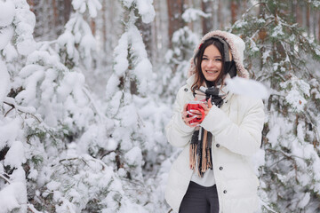Młoda ładna dziewczyna trzyma w dłoniach czerwony kubek z gorącą herbatą. Spaceruje w mroźny zimowy dzień po lesie. - obrazy, fototapety, plakaty