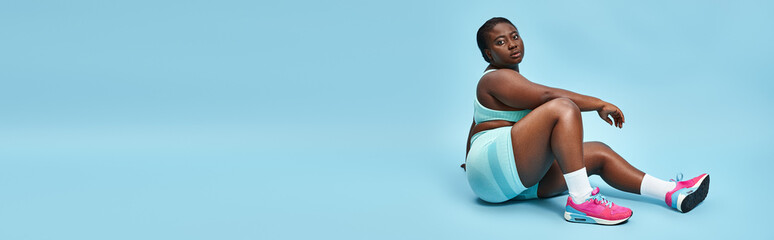 Relaxed plus size african american woman sitting in blue sportswear on matching backdrop, banner