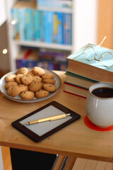 Cup of tea or coffee, pile of books, plate of cookies, reading glasses, e-reader and pen on the table. Colorful rainbow bookshelf in the background. Selective focus.