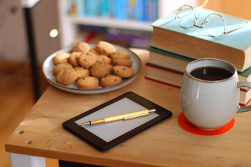 Cup of tea or coffee, pile of books, plate of cookies, reading glasses, e-reader and pen on the table. Colorful rainbow bookshelf in the background. Selective focus.