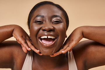 cheerful plus size african american woman smiling with hands near her face on beige background