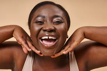 Fotobehang cheerful plus size african american woman smiling with hands near her face on beige background © LIGHTFIELD STUDIOS