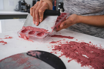 A woman holds in her hands a plastic mold filled with pink chocolate. A woman scrapes excess chocolate from a mold.
Kitchen work surface. Pink chocolate is poured into a plastic mold. 