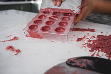 A woman holds in her hands a plastic mold filled with pink chocolate. A woman scrapes excess chocolate from a mold.
Kitchen work surface. Pink chocolate is poured into a plastic mold. 