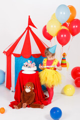 A little girl in a colorful outfit and a birthday hat is standing with a birthday party blower by...