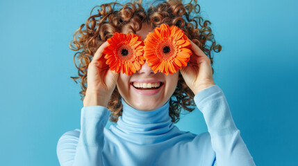 A person holds bright orange gerbera flowers over their eyes like glasses, smiling broadly against a blue background, creating a playful and joyful portrait.