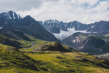 Dramatic scenery in alpine valley with creek among green hills and rocks with view to rocky sharp pointy peak, large snow-capped peaked top, mountain range and big glacier tongue under gray cloudy sky