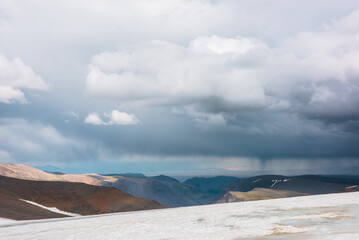 Dramatic alpine top view from flat sloping glacier on precipice edge to deep gorge between colorful sunlit cliffs and mountain range silhouette in rain under lead gray cloudy sky. Rainy grey clouds.