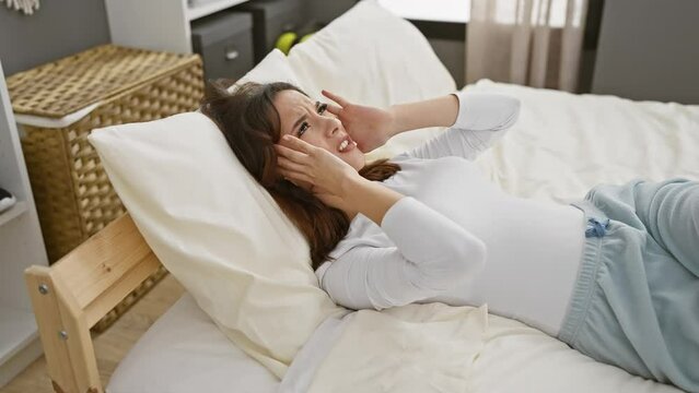 A young woman lies in bed covering her ears in frustration in a bright bedroom setting