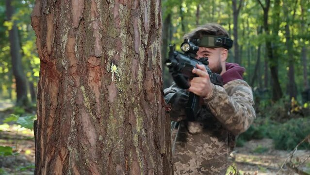 Laser tag, tree support, tactical gear. Man in camouflage attire and with marker on his head leans against tree, carefully aiming Laser tag gun and shoots at enemy in forest setting.