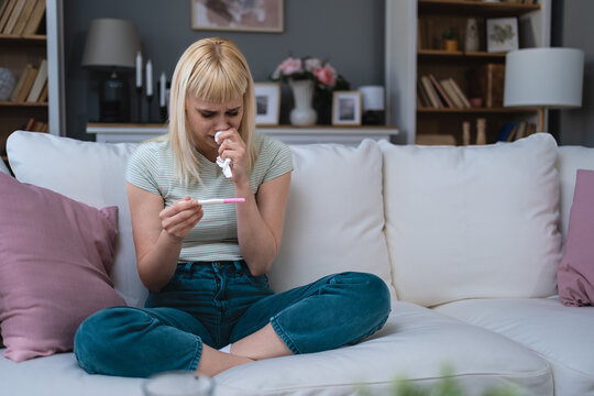 Single sad woman complaining holding a pregnancy test sitting on a couch in the living room at home, Stressed female, Positive or negative medical test pregnant or not.