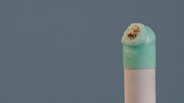 An ignited gelatin or agar pill of white and turquoise color, isolated on a grey background, burns, melts, foams, chars and turns into black lava mass under the influence of fire. The drugs and