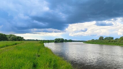 Shrubs and deciduous trees grow on the grassy banks of the river. Further away is the forest. A strong wind creates ripples on the water and drives clouds across the sky. Rain clouds gather