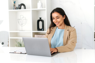 Work in the office. Happy indian or arabian business woman, company employee, wearing suit, using laptop at work, working on a project at workplace, searching information in internet, smiles