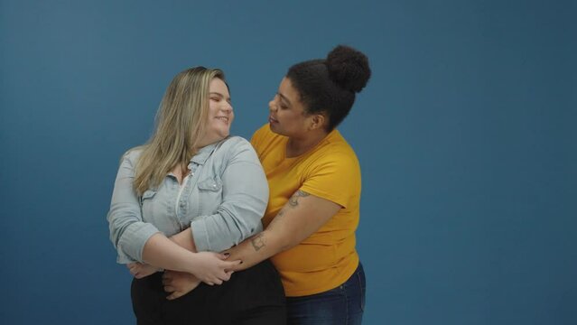 Sweet and tender kiss between two women in love shown of affection on isolated blue background studio. Free love to all sexual orientation. Gay young relationship.

