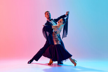 Elegant, passionate young couple, man and woman dancing ballroom in back stage costumes against gradient pink blue background in neon light. Concept of dance class, hobby, art, dance school, talent