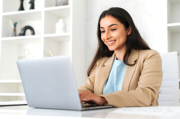 Working on a laptop. Positive successful indian or arabian business woman, company employee sitting at workplace in the office, using laptop, working on a project, looks at screen, smiling friendly