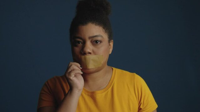 Black woman with sealed mouth sharply rip off sticky tape from lip. Don't be silent. Domestic and racism abuse and harassment concept. Isolated on blue background
