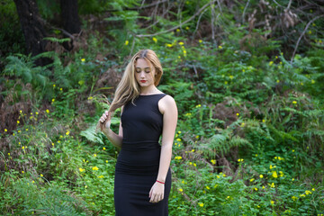 Young, beautiful blonde woman dressed in black walks through the forest in different postures and expressions. In the background ferns and yellow flowers. Concept expressions in nature.