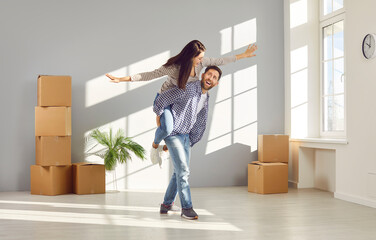 Young family couple having fun at home on moving day. Happy, cheerful man piggy backing his wife or...