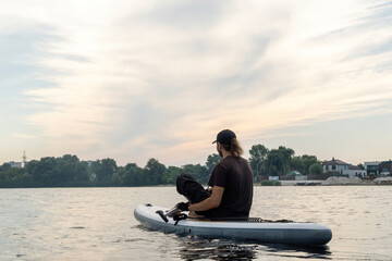 Young man sits on paddleboard amidst serene waters of village lake. Man enjoys active weekend and...